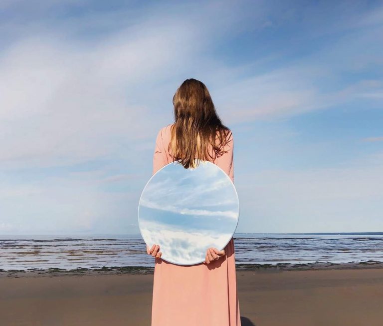 תמונת בחורה עומדת על החוף וצופה בים כשבידיה מראה עגולה שמתוכה משתקפים השמיים הכחולים- מתוך פוסט בנושא למה לשמור נגיעה באתר של מאור קפלן- מובילה תהליכי חינוך מיני במרחב הדתי